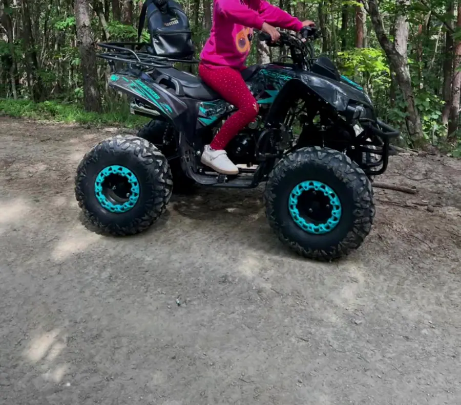 Fată în costum roz pe un ATV negru cu jante turcoaz, pe un drum de pădure