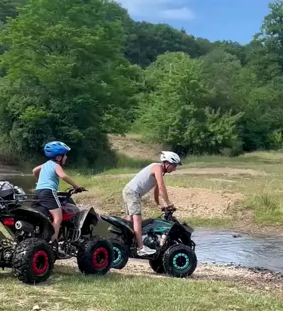 Doi copii cu căști trec cu ATV-urile pe lângă un pârâu
